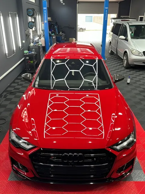 Rotes Auto frontal in einer Aufbereitungshalle, das sechseckige Deckenlicht spiegelt sich als Wabenmuster auf der Motorhaube.