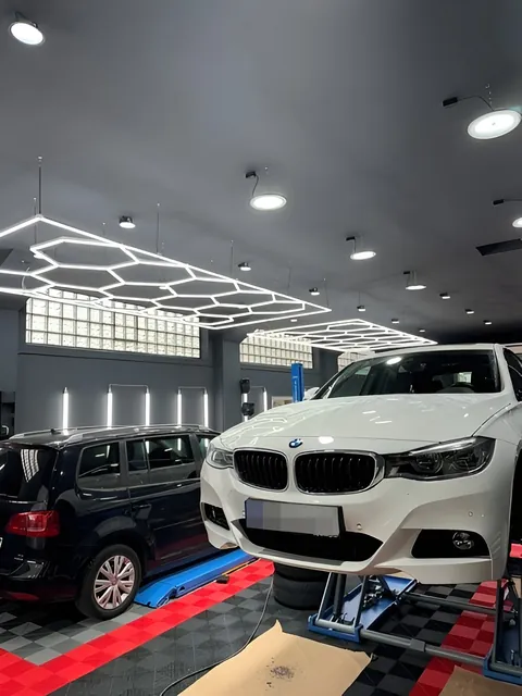 Weißes Auto frontal auf einer Hebebühne, links ein schwarzer Transporter, Strahler an der Decke.